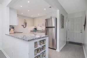 A modern kitchen with white cabinetry, a stainless steel refrigerator, granite countertops, and a decorative wall piece. - Pet Friendly Townhome Directly Across from the Beach in Quiet Ormond By the Sea (Ormond Beach)