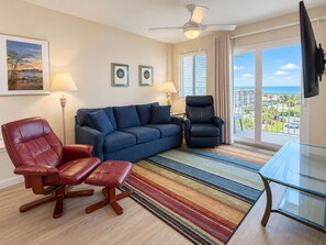 Ein Wohnzimmer mit einer Couch, zwei Sesseln, einem Glastisch, einem Deckenventilator und Blick auf den Ozean durch große Fenster.
