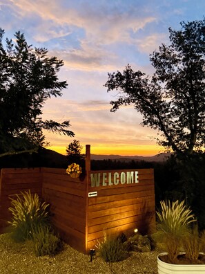 A wooden welcome sign with a directional arrow, surrounded by plants and trees, with a sunset in the background.