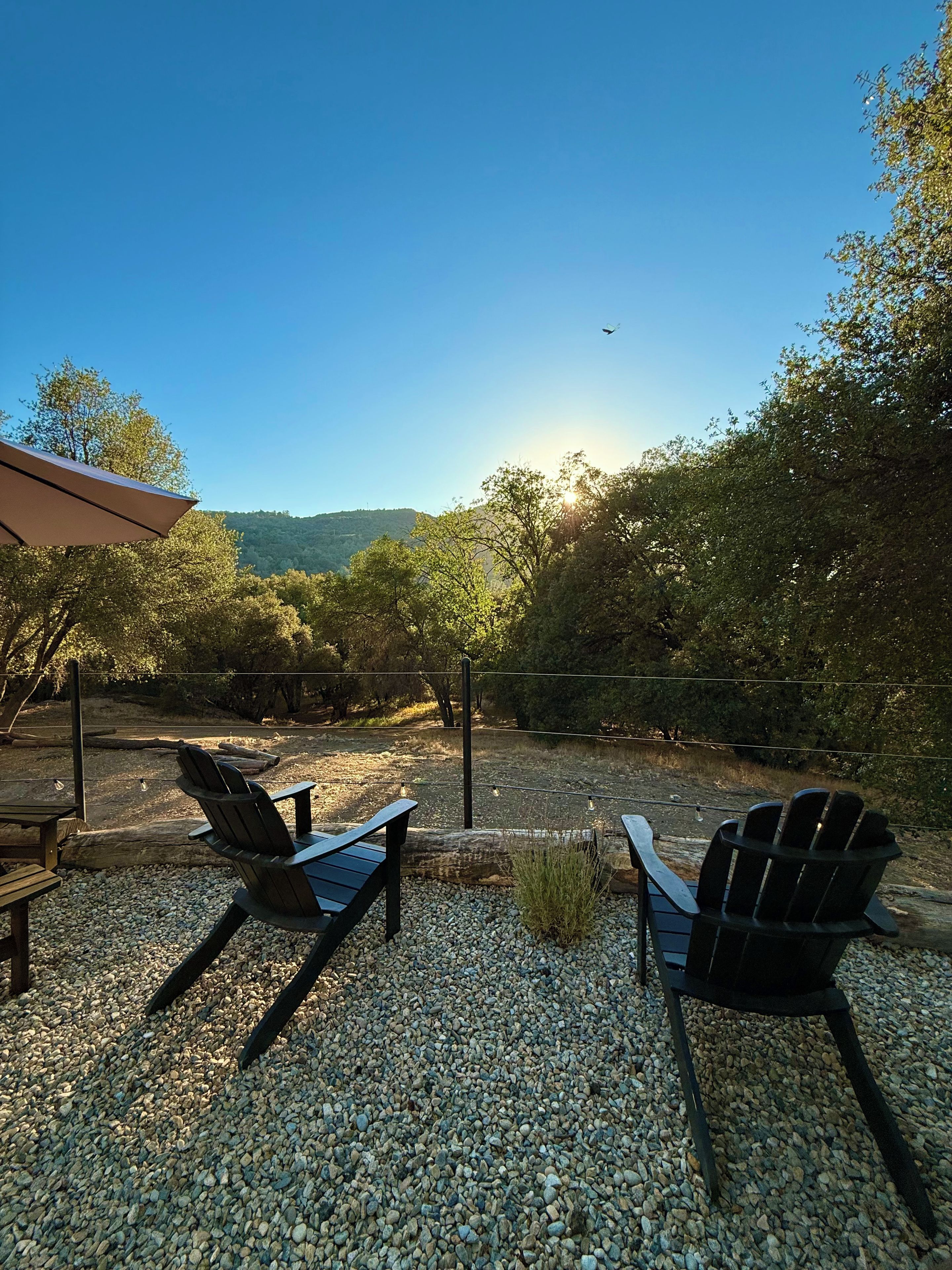 An outdoor seating area with Adirondack chairs, a wooden table, and a gravel ground.