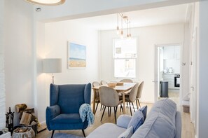A modern living room with a blue sofa, a dining area with a table and chairs, a kitchen in the background, and a beach-themed painting on the wall. - 🏖️ Seaside Charm in Broadstairs – Cosy 2-Bed Home with Private Parking (Broadstairs)