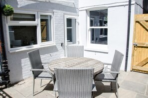 En terrasse med et rundt bord og fire fletstole, hvide vægge og en trædør.