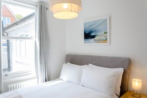 A bedroom with a bed, a nightstand, a lamp, a window with curtains, and a painting on the wall. - 🏖️ Seaside Charm in Broadstairs – Cosy 2-Bed Home with Private Parking (Broadstairs)