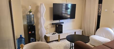 A modern living room with a flat-screen TV mounted on the wall, a water dispenser, a white vase, a small table with a smartphone, and a sofa.