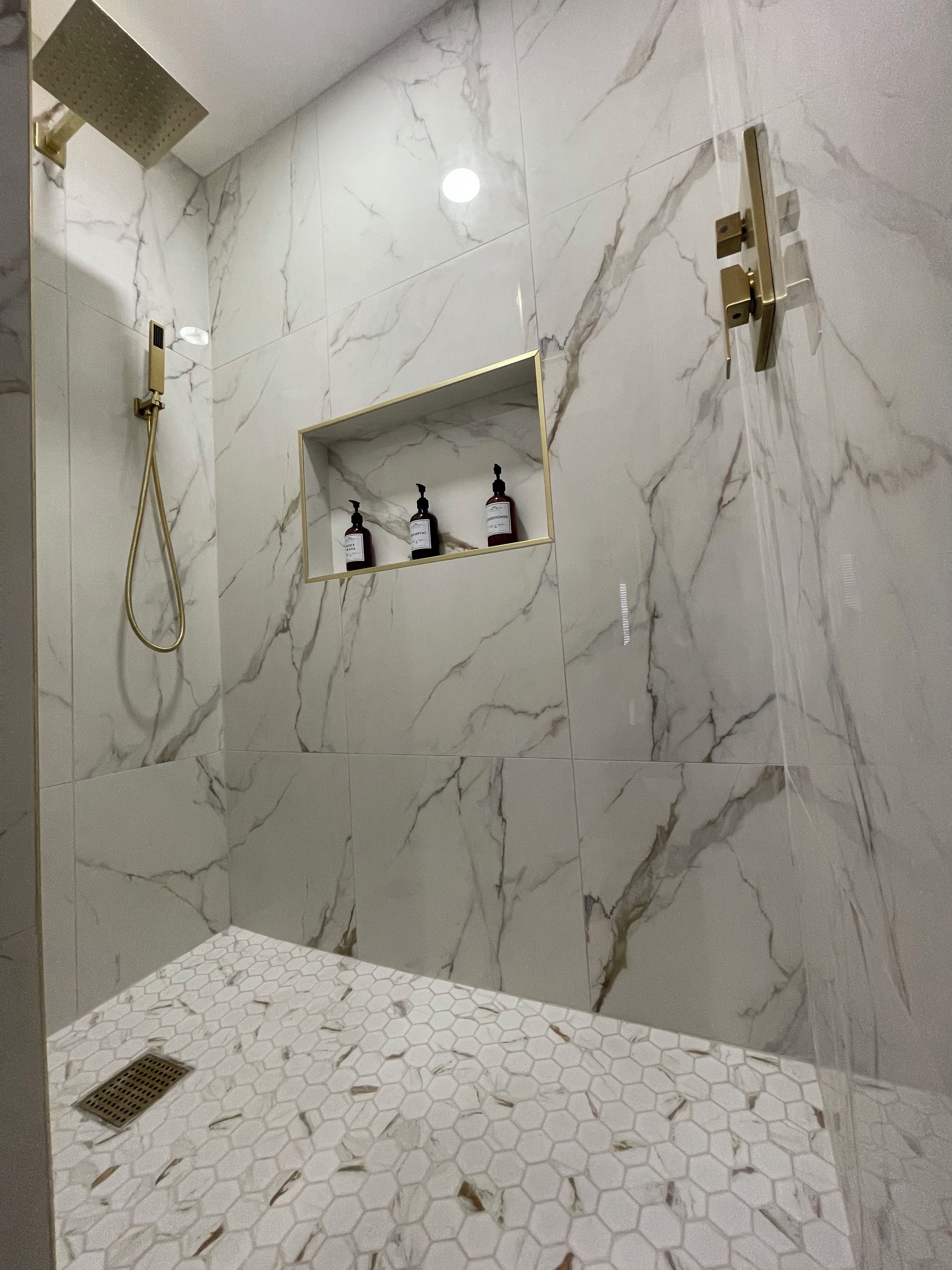A bathroom with marble walls, a shower with a handheld showerhead, and a shelf with toiletries.