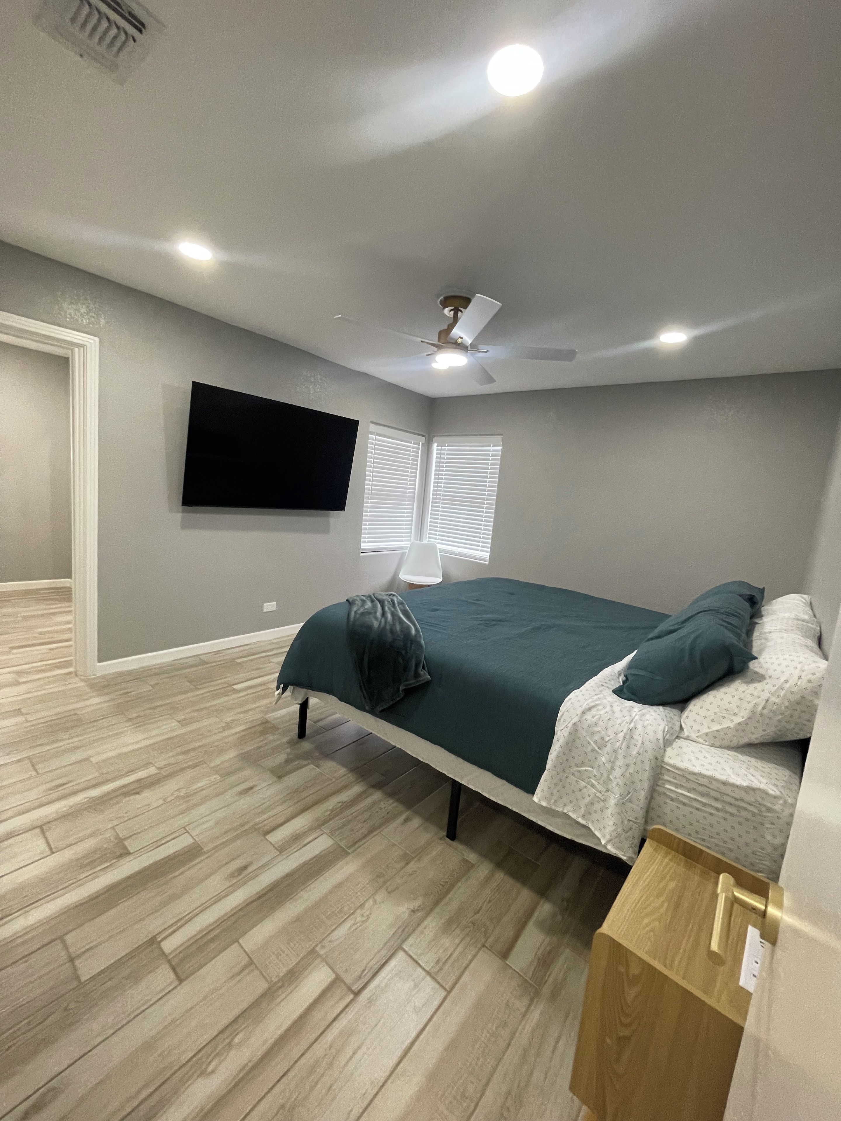 Une chambre à coucher avec un lit, un ventilateur de plafond, un téléviseur fixé au mur et une fenêtre avec des stores.