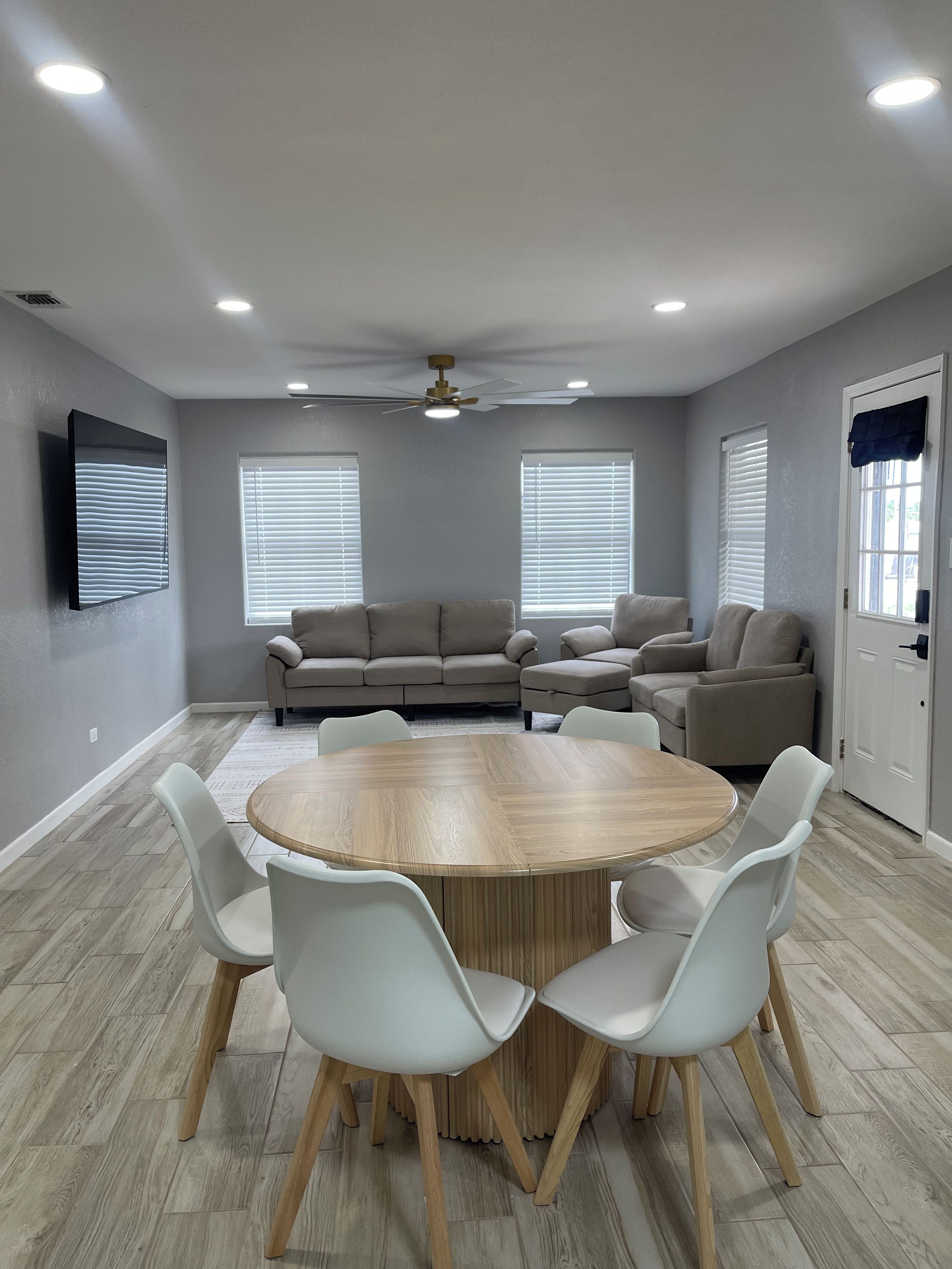 Une pièce avec une table ronde en bois, entourée de chaises blanches, un canapé gris et un ventilateur de plafond.
