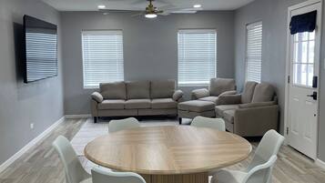 Une pièce avec une table ronde en bois, entourée de chaises blanches, un canapé gris et un ventilateur de plafond.