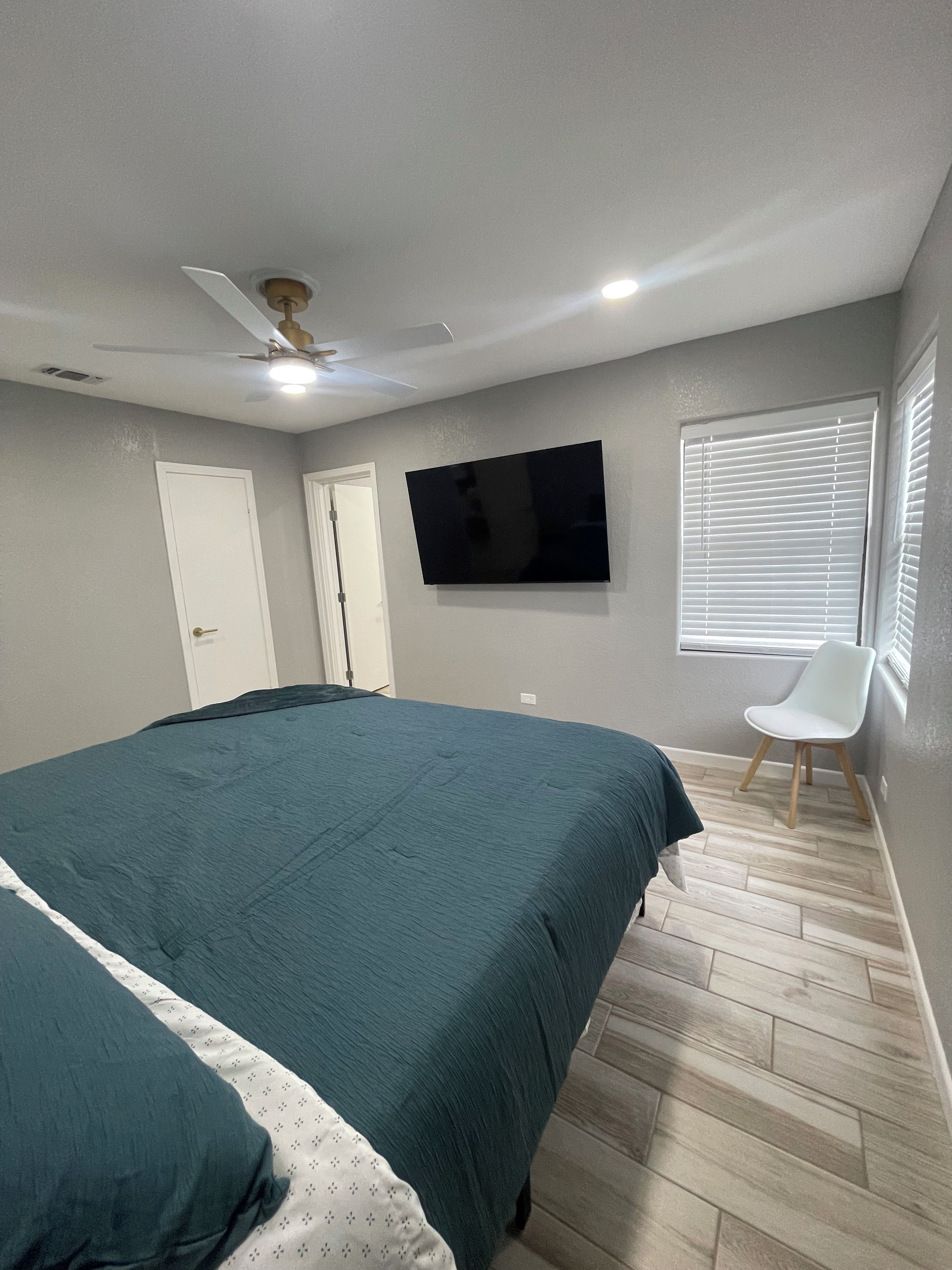 A bedroom with a bed, a television, a ceiling fan, a window with blinds, and a chair.