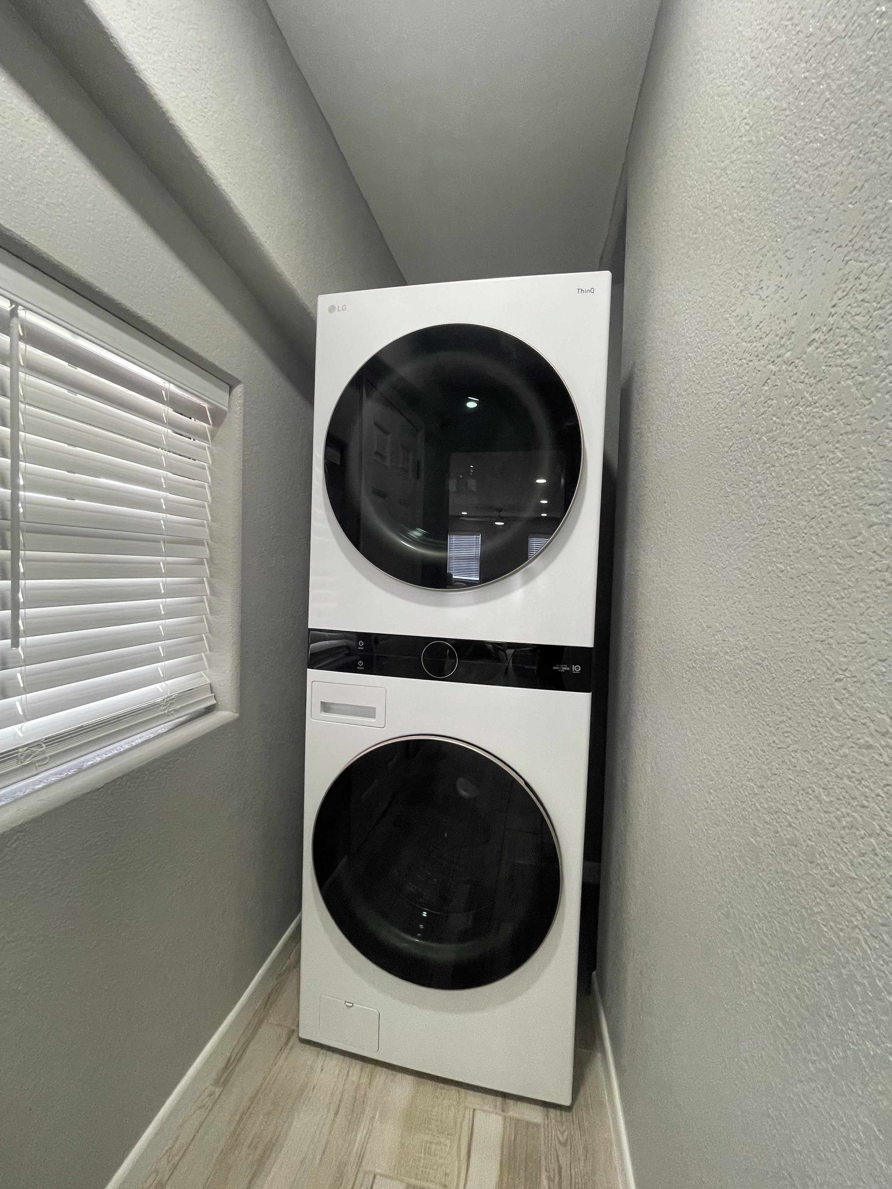 A compact, white, wall-mounted washer and dryer unit in a narrow hallway with a window with blinds on the left.