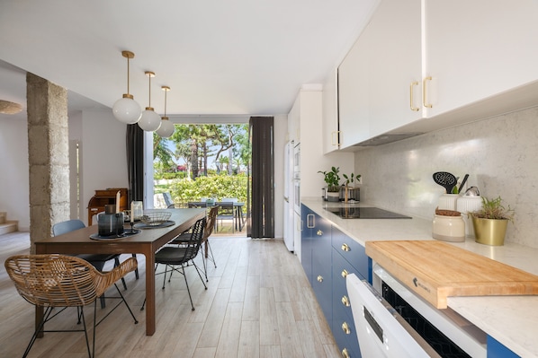 A modern kitchen with a dining area, featuring a wooden table, chairs, and a view of a garden through the window.