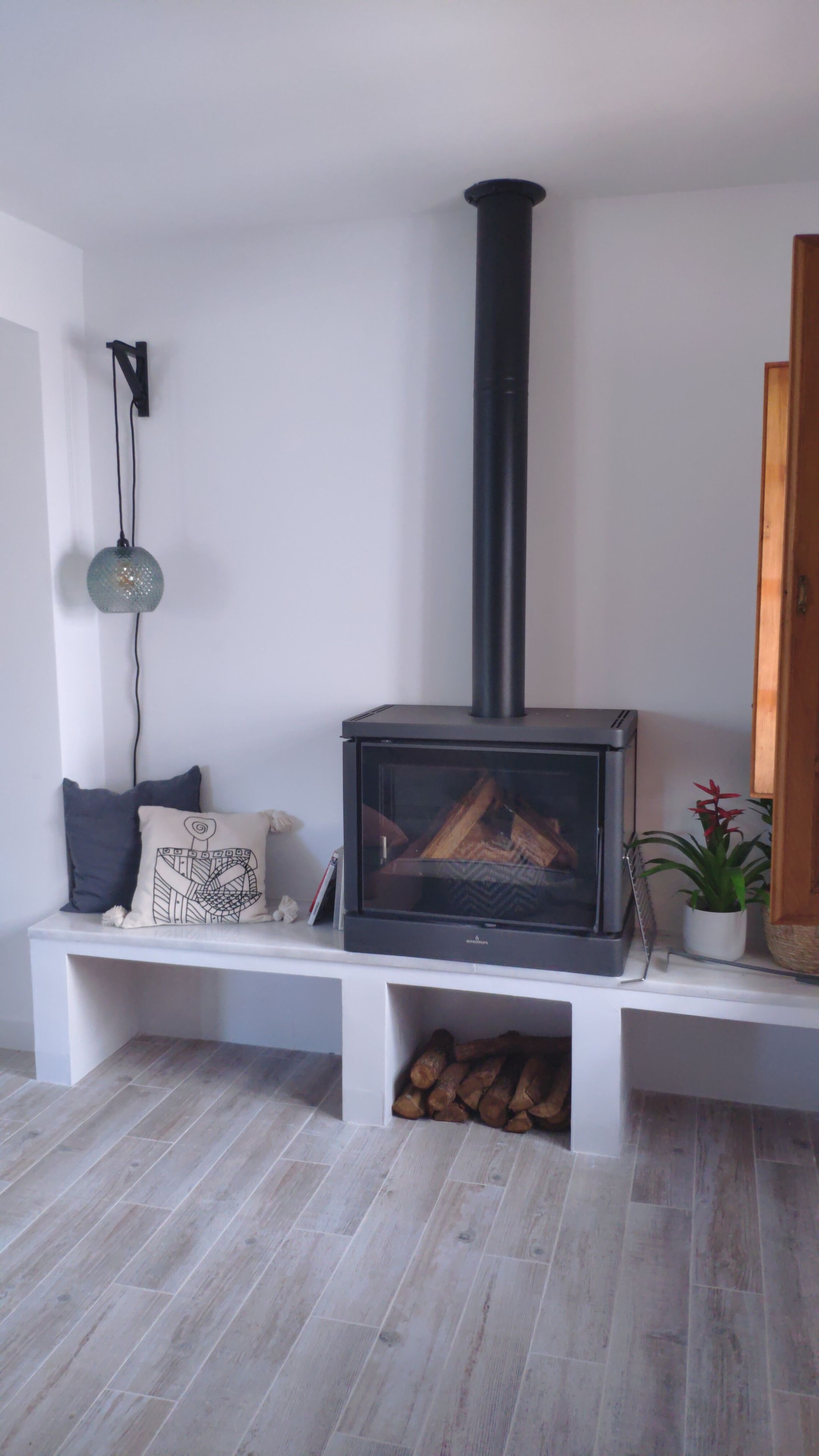 Un salon moderne doté d’un poêle à bois encastré, d’un banc avec des coussins, d’une lampe suspendue et d’une plante en pot.