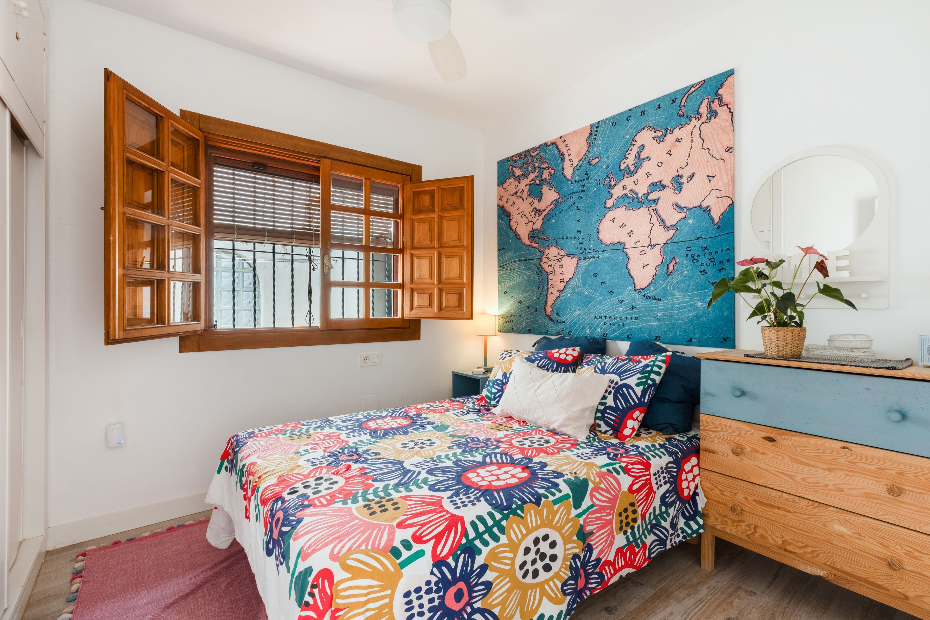 A bedroom with a wooden window, a bed with a colorful floral bedspread, a world map on the wall, and a dresser with a mirror.