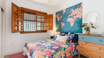 A bedroom with a wooden window, a bed with a colorful floral bedspread, a world map on the wall, and a dresser with a mirror.