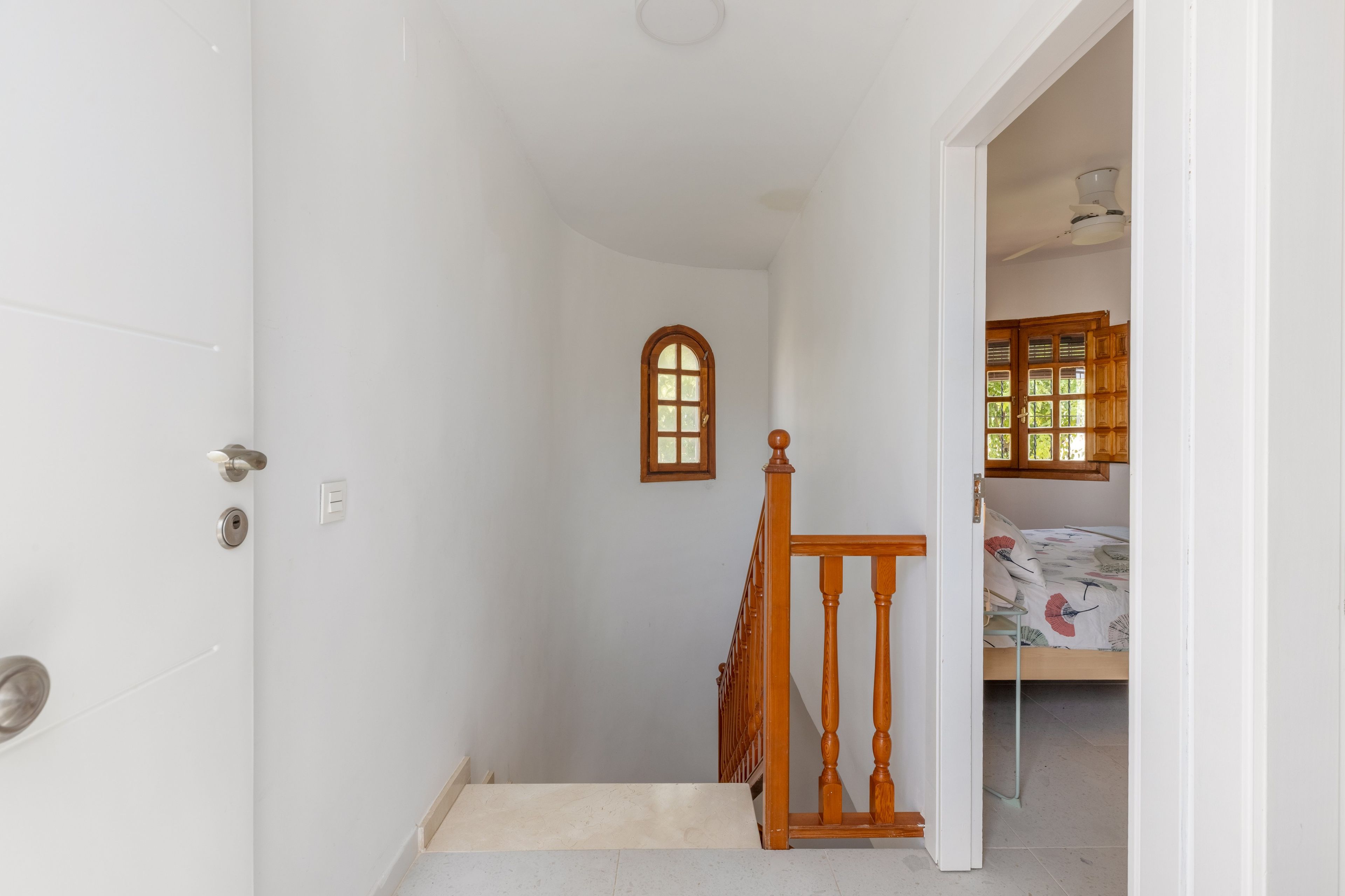 A narrow hallway with a wooden door, a small window, and a staircase leading to a bedroom with a bed and a window.
