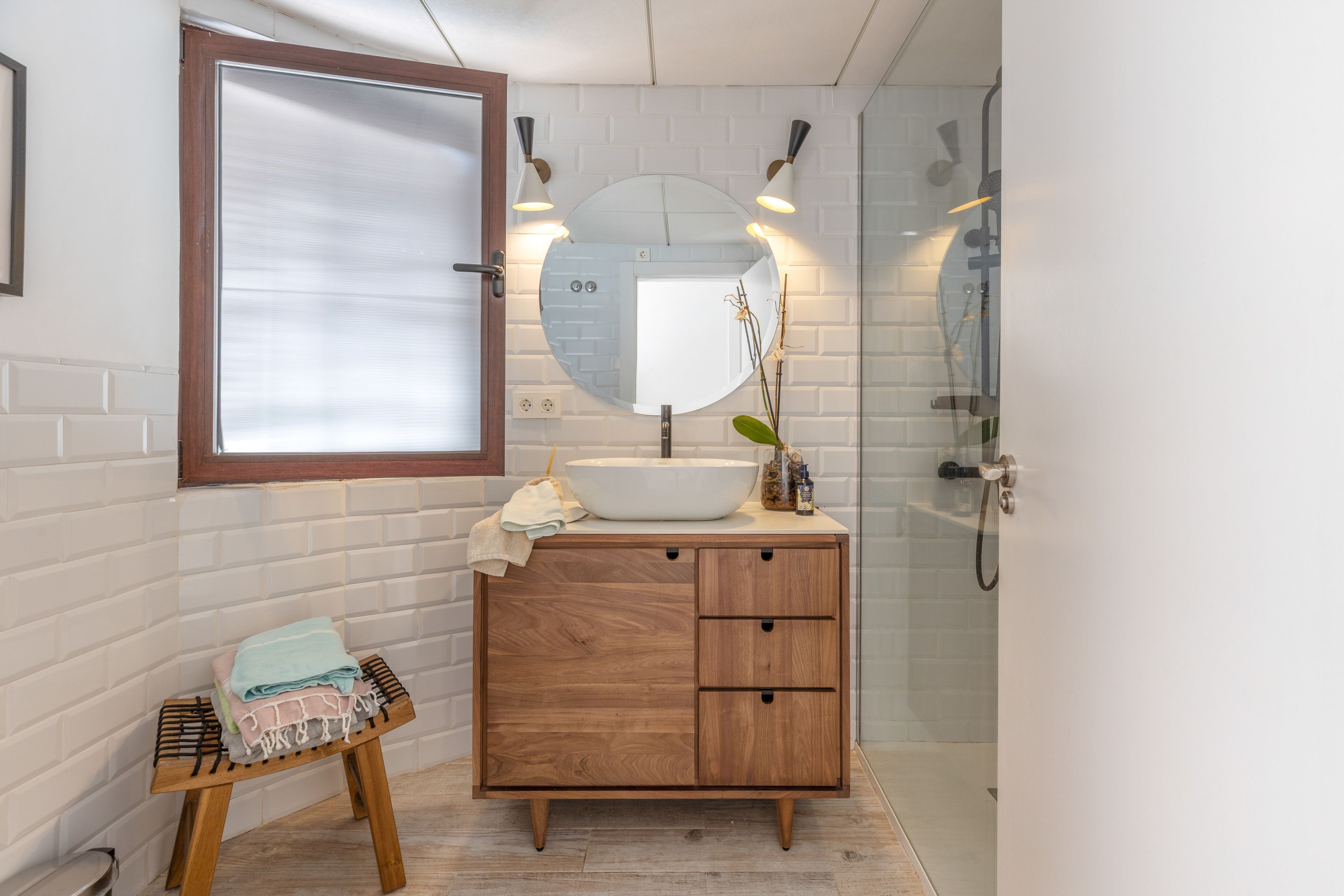 Un baño con tocador de madera, espejo redondo y cabina de ducha con mampara de vidrio.