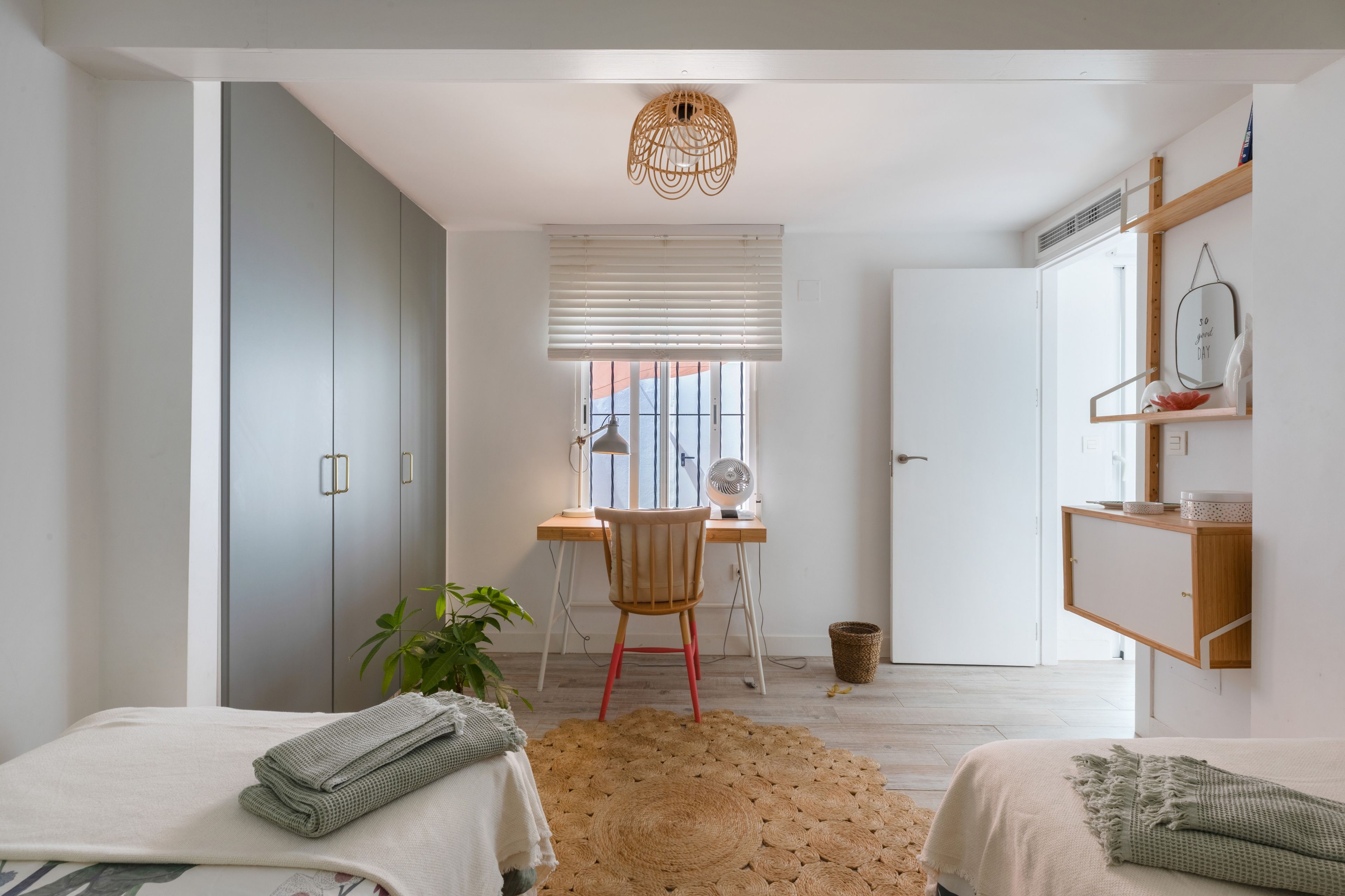A bedroom with a desk, two beds, a hanging light fixture, and a view of the outside.