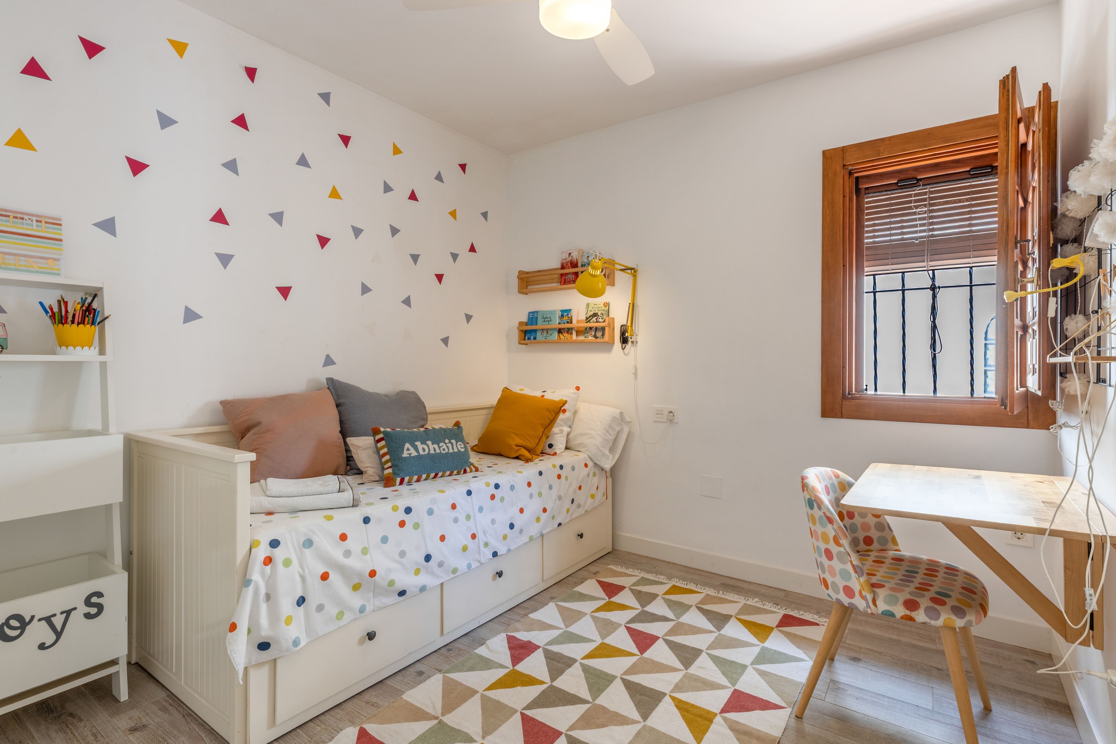 Una habitación infantil con una cama blanca, colcha con lunares de colores, un escritorio pequeño con silla, una estantería y un muro con adornos geométricos.