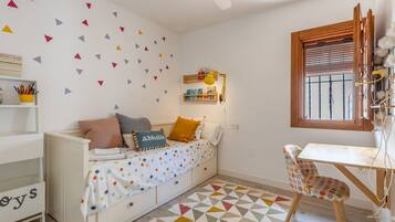 Una habitación infantil con una cama blanca, colcha con lunares de colores, un escritorio pequeño con silla, una estantería y un muro con adornos geométricos.