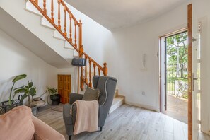 Ein geräumiges Wohnzimmer mit einem grauen Sessel, einer Holztreppe und Blick auf einen Garten durch eine offene Tür.