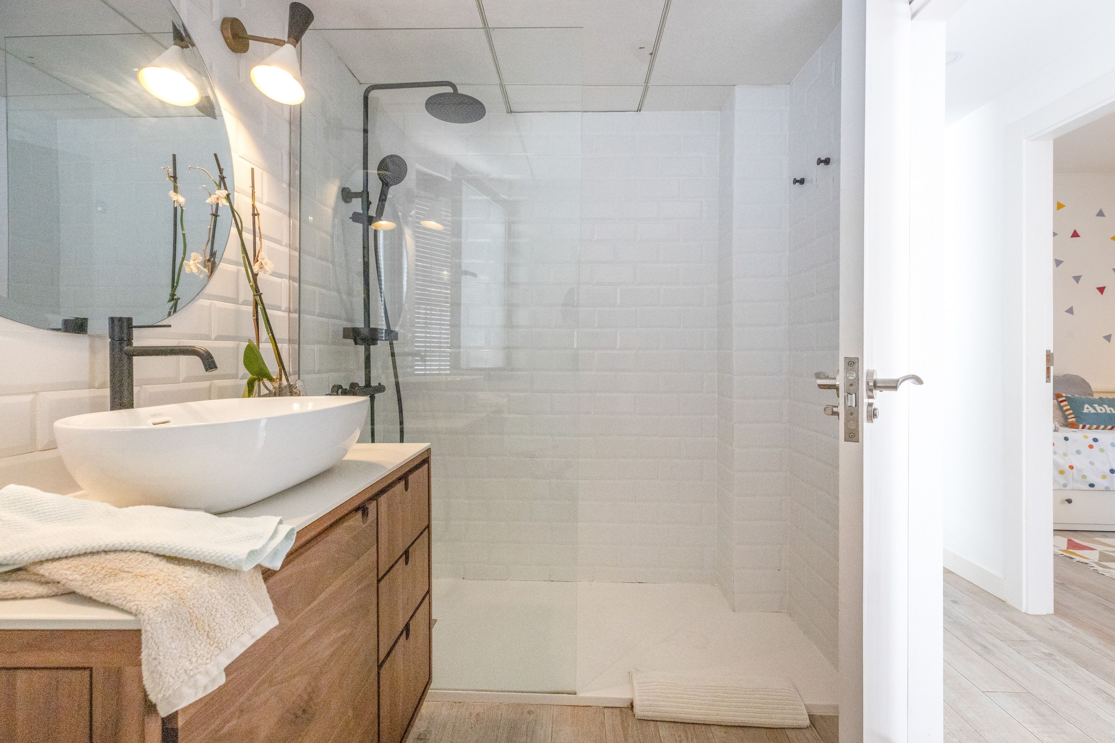 Un baño con lavamanos blanco, espejo y ducha. Hay una toalla en el suelo y un armario de madera.