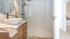 A bathroom with a white sink, mirror, and shower. There is a towel on the floor and a wooden cabinet.