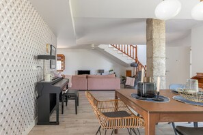 A modern living room with a piano, a dining table with chairs, a staircase, and a large pillar.