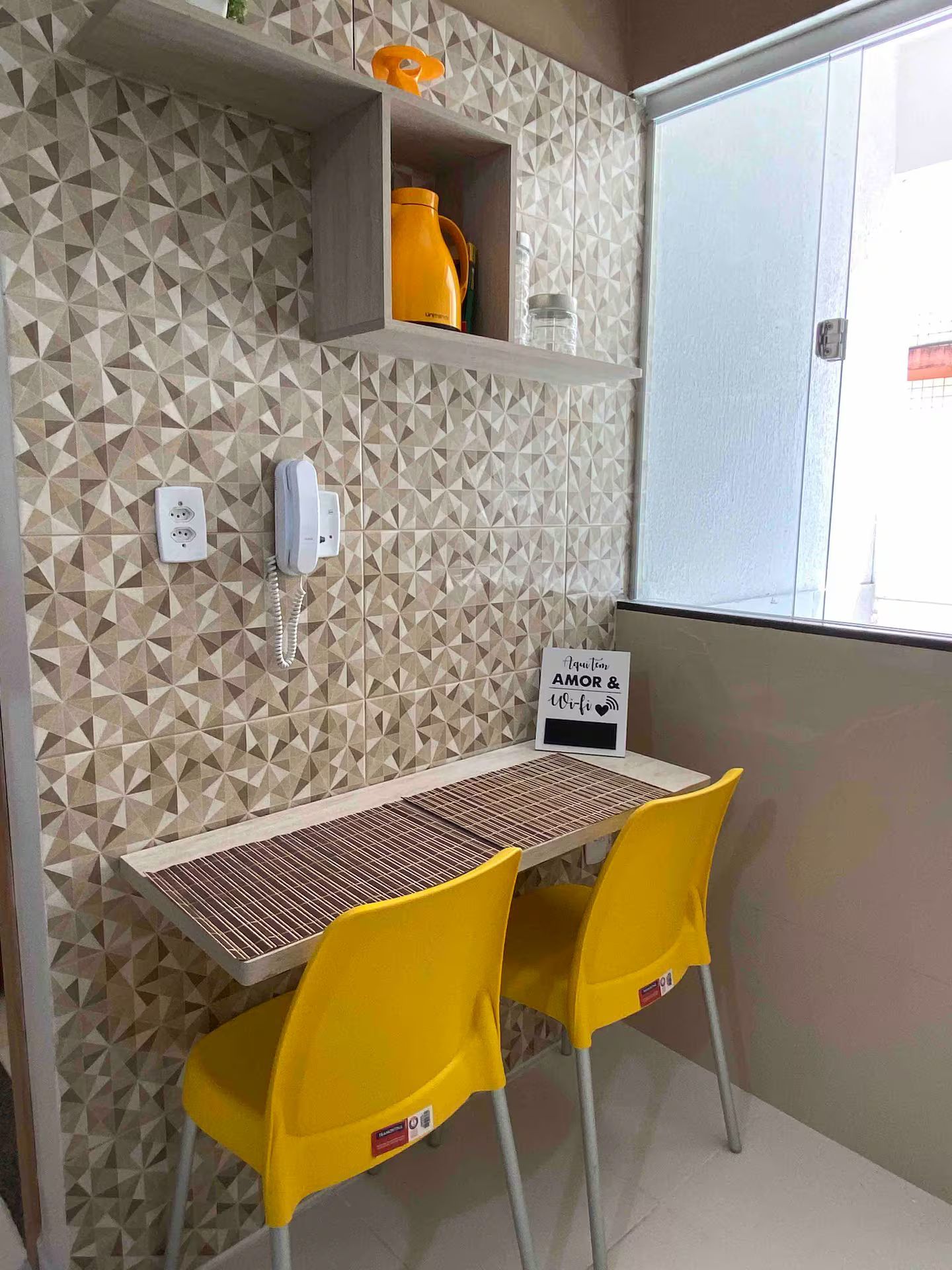 A small room with a geometric patterned wall, a table with two yellow chairs, a whiteboard with a message, and a window with a view.