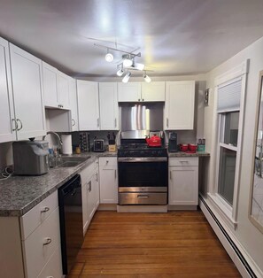 A compact kitchen with white cabinets, a stainless steel range, a dishwasher, and a window with blinds.