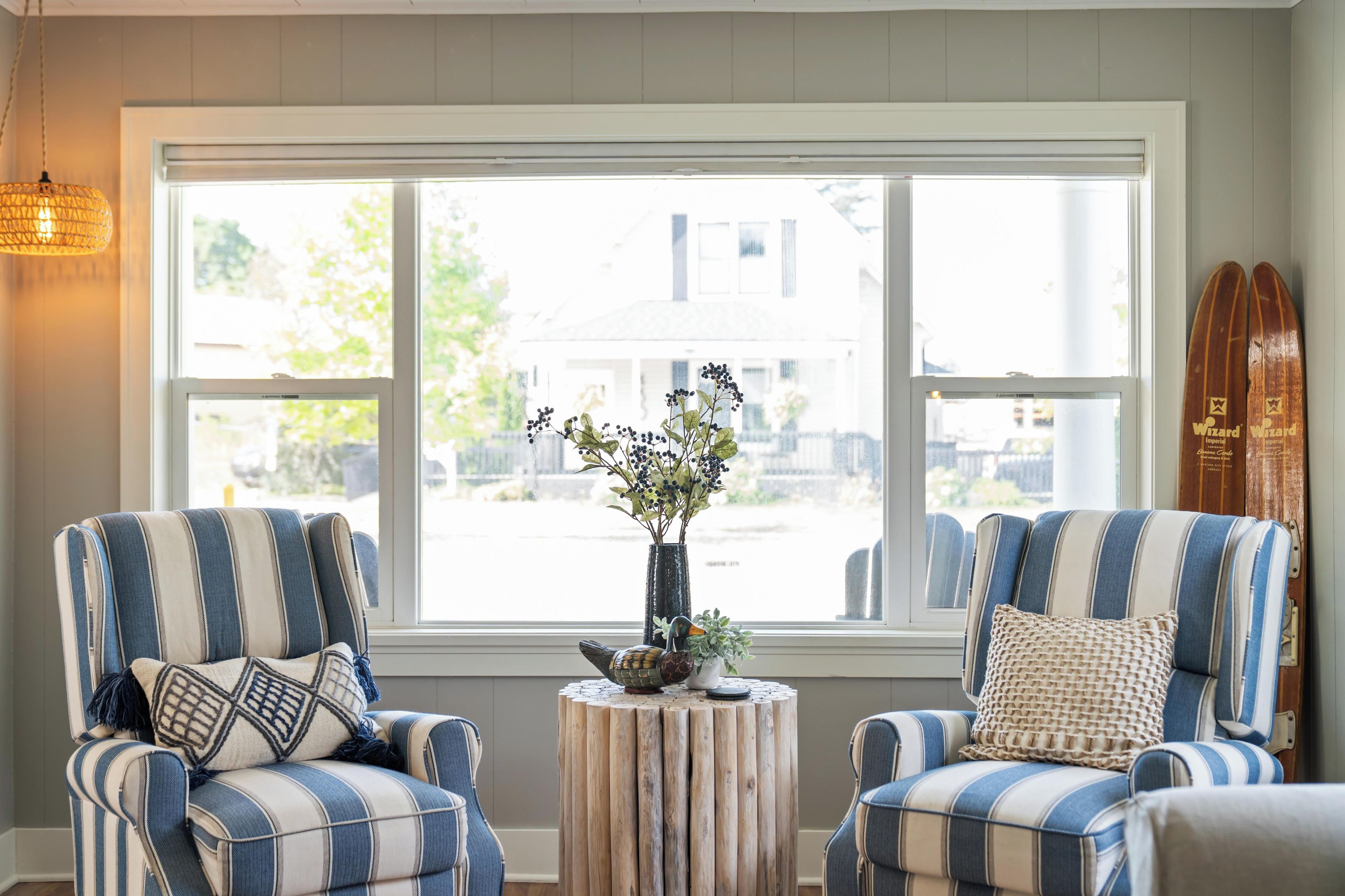 A room with two striped armchairs, a small wooden table with a vase of flowers, and a large window with a view of a building outside.