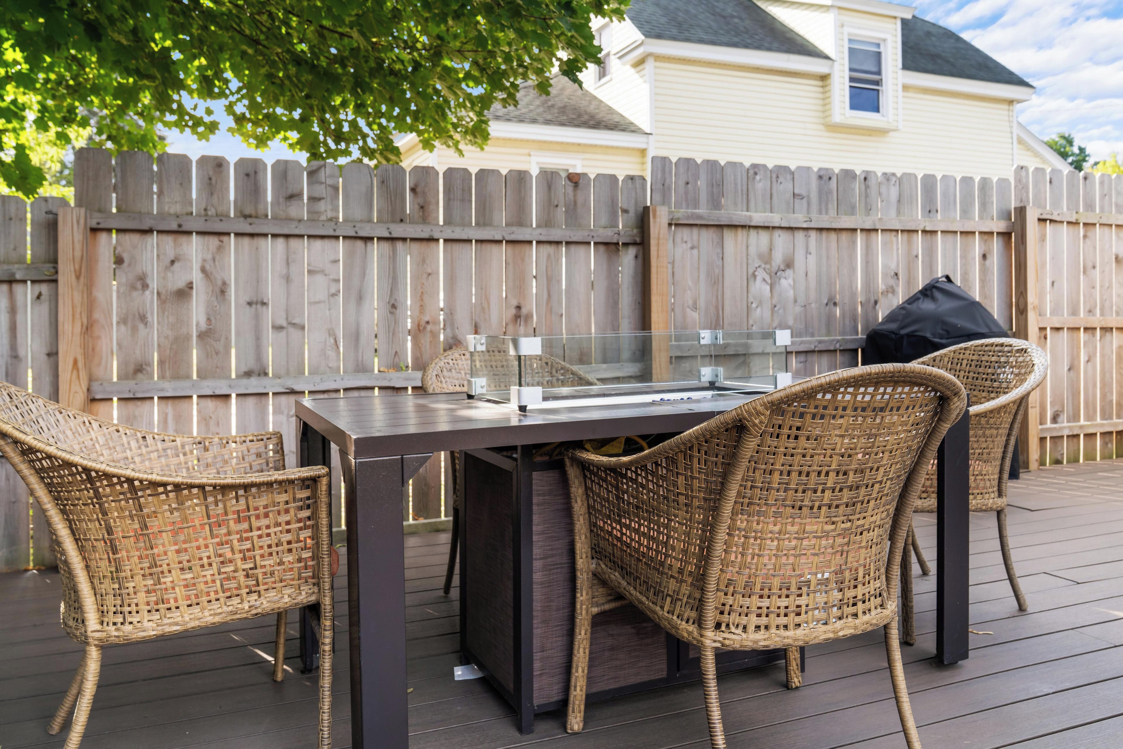 A patio with a table and wicker chairs, a glass-top table, and a wooden fence in the background.
