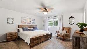 A bedroom with a wooden bed, a ceiling fan, a window with curtains, a chair, a dresser, and wall art. - Country Club - Large Yard - Modern & Spacious (Gainesville)