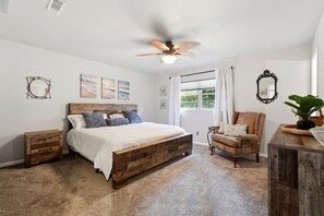 Ein Schlafzimmer mit einem hölzernen Bett, Deckenventilator, einem Fenster mit Vorhängen, einem Sessel, einem Schminktisch und Wandbildern.
