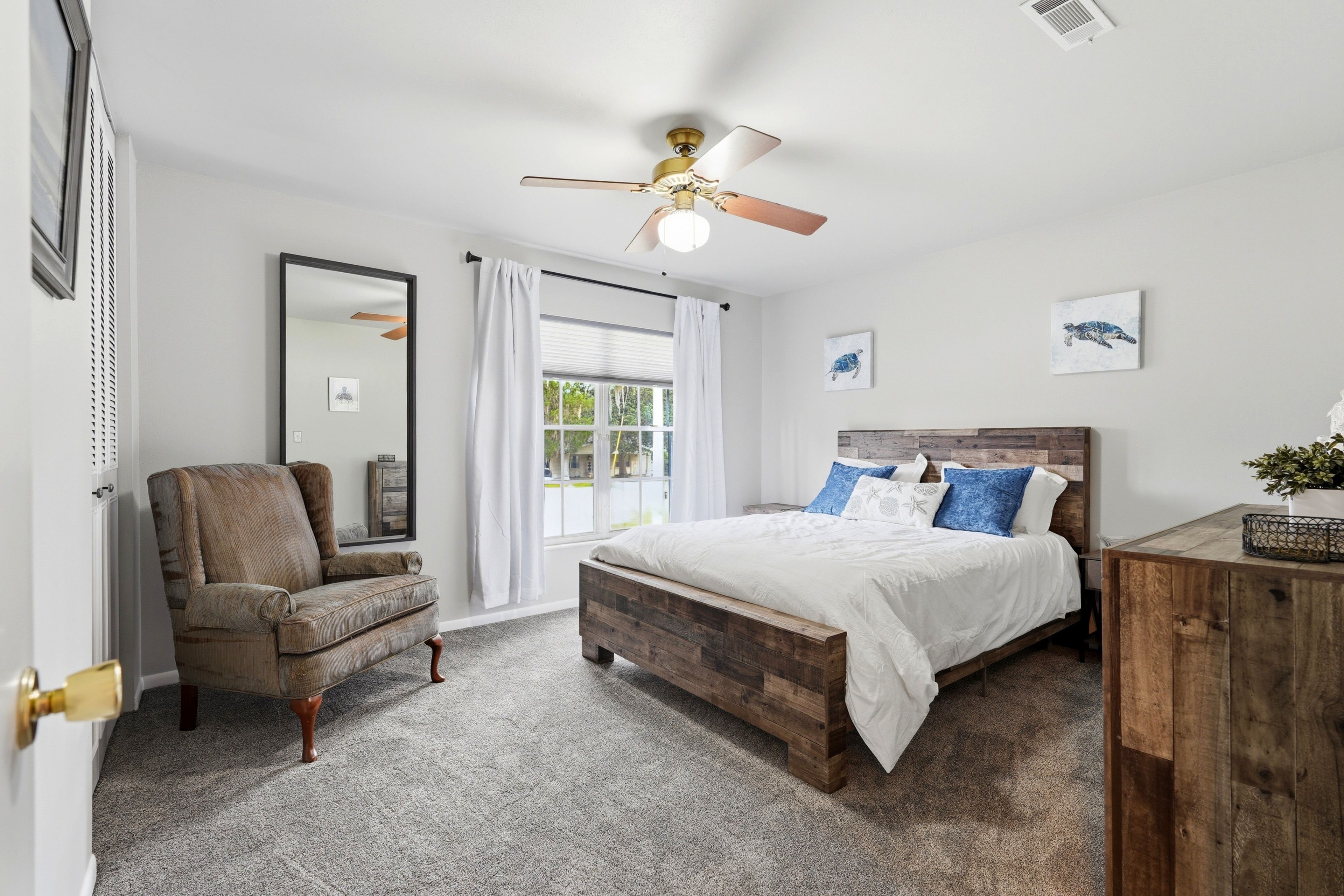 A bedroom with a wooden bed, a chair, a ceiling fan, and a window with curtains.