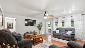 Ein Wohnzimmer mit Deckenventilator, Flachbildfernseher, grauen Sofas, einem Holztisch und einem großen Fenster mit durchsichtigen Vorhängen.