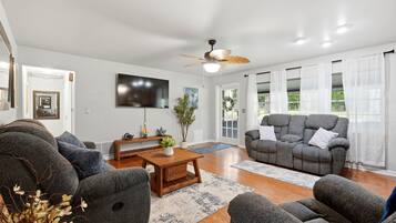 A living room with a ceiling fan, a flat-screen TV, grey sofas, a wooden coffee table, and a large window with sheer curtains.