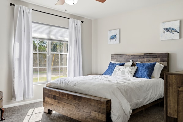 Un dormitorio con una cama de madera, ropa de cama blanca y vistas al exterior a través de un gran ventanal con cortinas blancas.