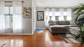 Ein Wohnzimmer mit einem grauen Sofa, einem Couchtisch, einer Lampe und einem großen Fenster mit weißen Vorhängen.