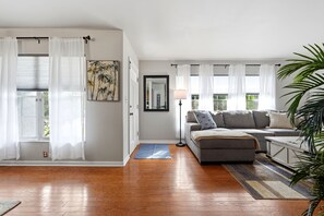 Una sala de estar con un sofá gris, una mesita de centro, una lámpara y un ventanal con cortinas blancas.