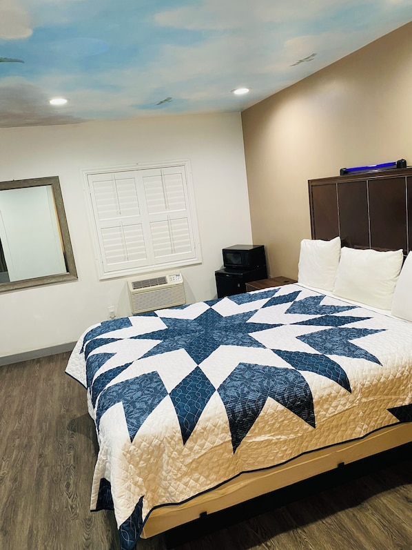 Une chambre à coucher comprenant un lit, une table de chevet, une commode, une fenêtre avec des stores et un plafond décoré d’un motif de ciel peint.
