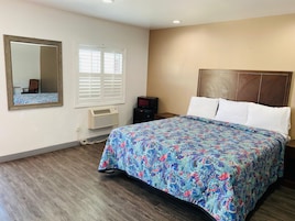 Habitación de hotel con cama, un espejo grande, una ventana con persianas, un equipo de aire acondicionado y un televisor.