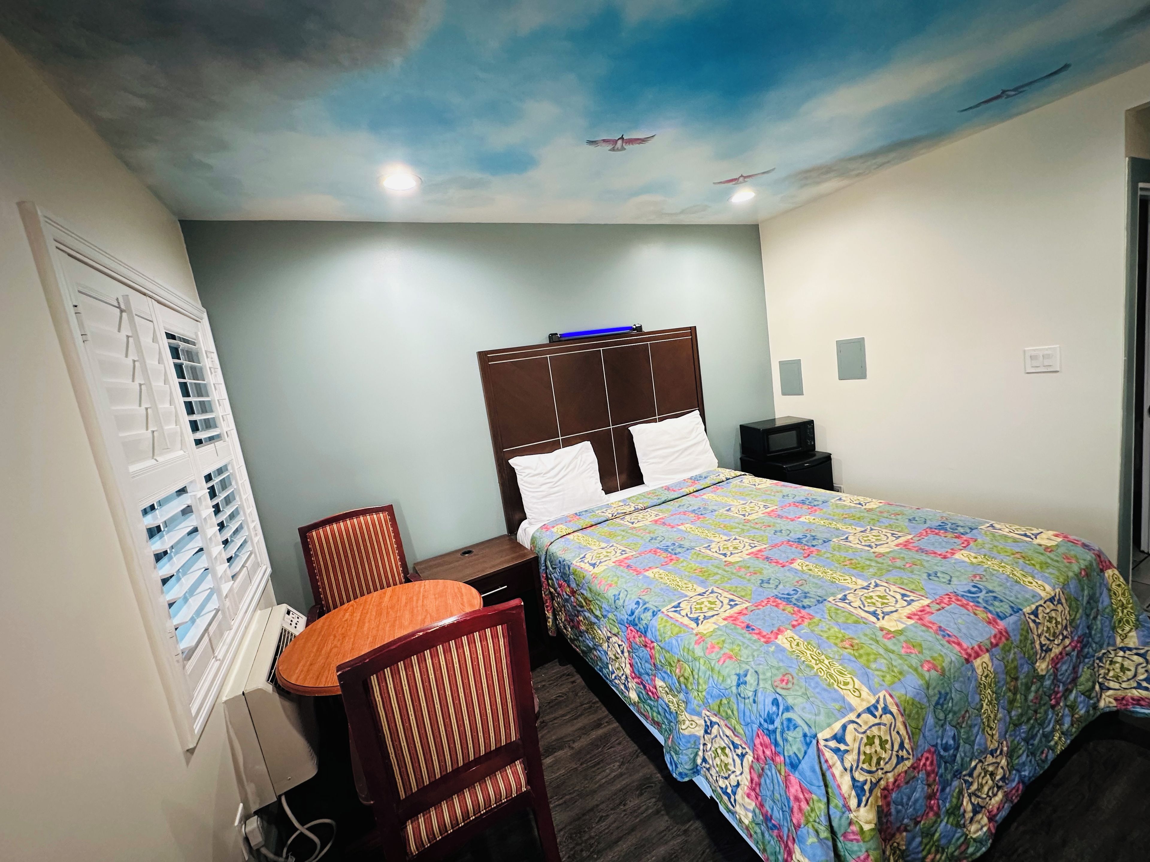 A hotel room with a bed, a desk, a chair, a television, and a ceiling mural.