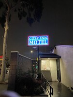 Un letrero de motel con contorno de neón, una entrada con portones y un pequeño edificio con una puerta visible.