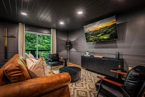 Ein modernes Wohnzimmer mit einer Ledersofa, einem schwarzen Sessel, einem kleinen runden Tisch, einer schwarzen Kommode und einem großen Fernseher an der Wand, der eine malerische Landschaft zeigt.