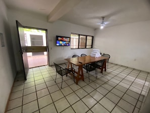 Ein Zimmer mit Fliesenboden, einem Holztisch mit Stühlen, einem an der Wand montierten Fernseher und einem Deckenventilator.