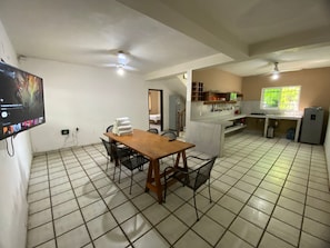 Ein Zimmer mit Fliesenboden, einem Holzespeisetisch mit Stühlen, einem an der Wand montierten Fernseher und einer Küchenzeile mit Kühlschrank und Fenster.