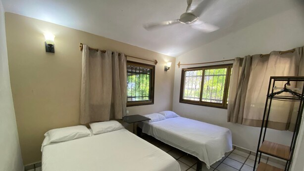 A room with two beds, a ceiling fan, a window with curtains, and a clothing rack. - Tzalahua House (Manzanillo)