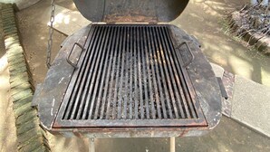 A large, open metal grill with a chain attached, set on a concrete surface with a white door and a plant in the background. - Tzalahua House (Manzanillo)