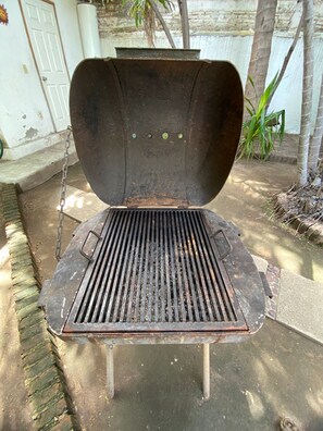 Ein großer, offener Metallgrill mit einer daran befestigten Kette, auf einer Betonfläche mit einer weißen Tür und einer Pflanze im Hintergrund.