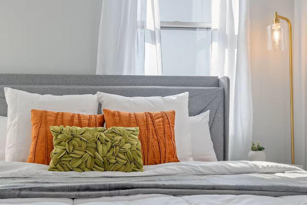 A neatly made bed with a grey headboard, white and orange pillows, and a green leaf-patterned pillow.